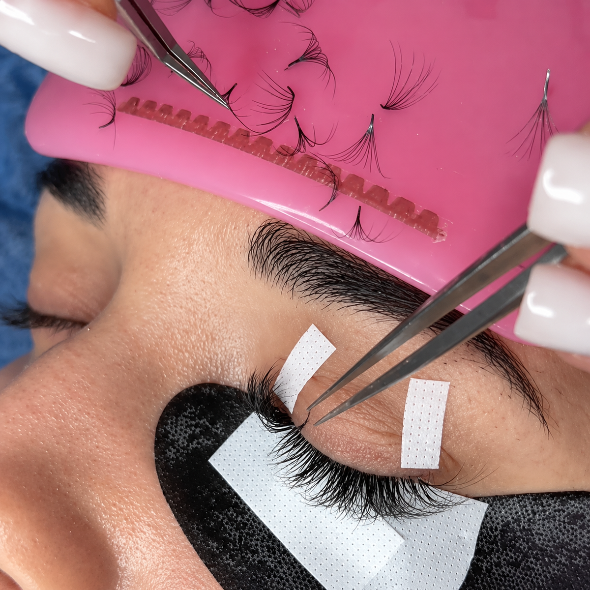 Close-up of a lash artist applying eyelash extensions using tweezers with under-eye pads and lash strips on a pink lash holder.