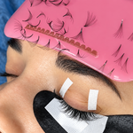 Close-up of eyelash extensions being applied with under-eye pads and lash strips displayed on a pink lash holder.