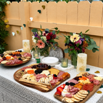 Outdoor brunch setup with a charcuterie board, fruits, cheese, and floral decorations.
