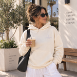 Person wearing a beige oversized Thousand Lashes hoodie with a front logo while holding a coffee outdoors.