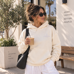 Person wearing a beige oversized Thousand Lashes hoodie with a front logo while holding a coffee outdoors.
