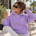 Person wearing a lavender oversized Thousand Lashes hoodie with a front logo while sitting outdoors and holding a coffee cup.