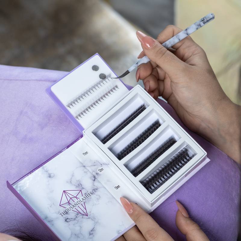 Lash artist holding a marble-pattern tweezer above a lash extension tray with multiple lash styles arranged in a case.