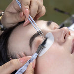 Lash artist using tweezers to apply eyelash extensions while a client rests with under-eye gel pads in place.